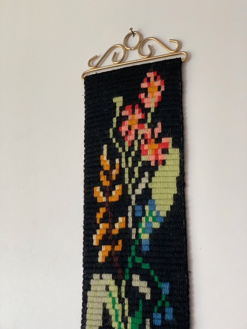 May include: A black tapestry with a floral design in shades of orange, pink, green, and blue. The tapestry is rectangular and hangs from a gold-colored metal bracket. The flowers are stylized and arranged in a vertical pattern.