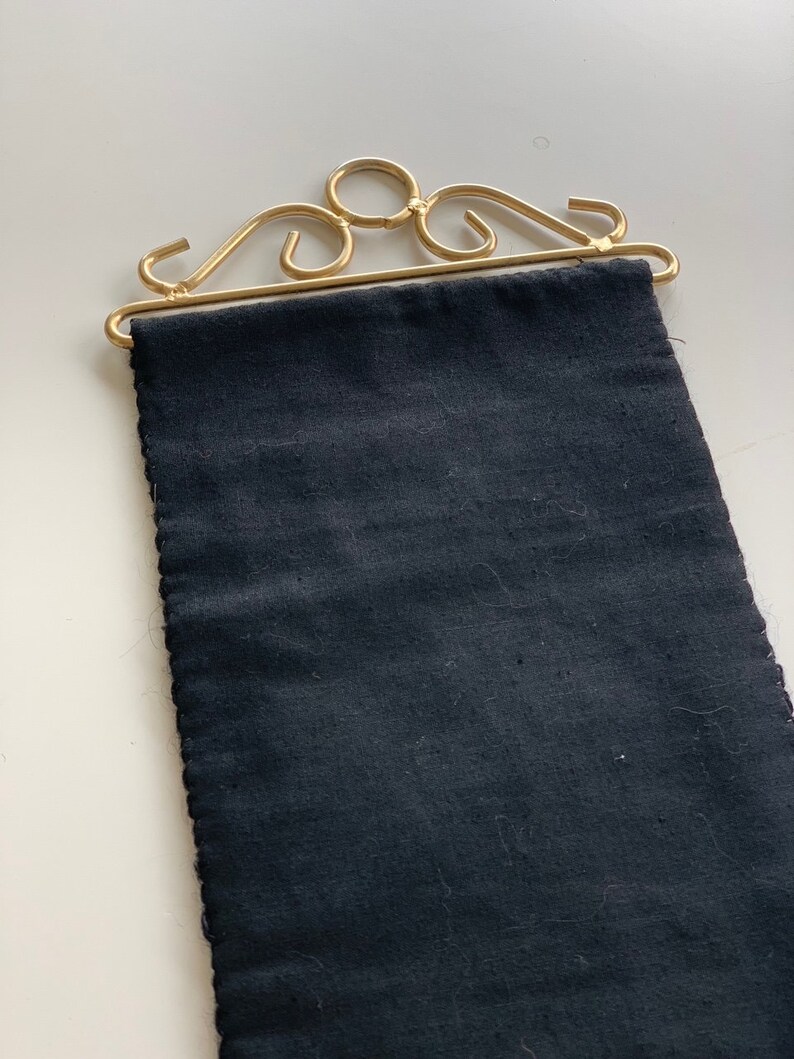 May include: A decorative wall hanging featuring a dark blue fabric panel suspended from a gold-colored metal frame. The frame has an ornate, scrolled design. The fabric has a textured appearance.