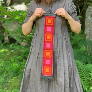 Könnte beinhalten: Ein rechteckiger Wandbehang mit gehäkelten Quadraten in Rosa, Orange und Rot. Die Quadrate sind von einem dekorativen, dunklen Metallrahmen eingefasst. Der Hintergrund ist eine verschwommene Außenszene mit grünem Laub. Der Behang ist ca. 60 cm lang.