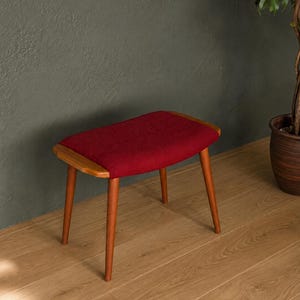 May include: A small stool featuring a rectangular seat upholstered in deep red fabric. The frame is constructed from light brown wood, showcasing four tapered legs that extend outward from the base to support the cushioned seat top.