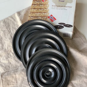 Op de afbeelding: Vier zwarte metalen taartringen voor het maken van een traditionele Noorse kransekake-taart. De ringen zijn in elkaar geschoven en hebben een antiaanbaklaag. De verpakking toont een afbeelding van een afgewerkte kransekake-taart en de tekst "kransekakeformer", wat "kransekake-vormen" betekent.