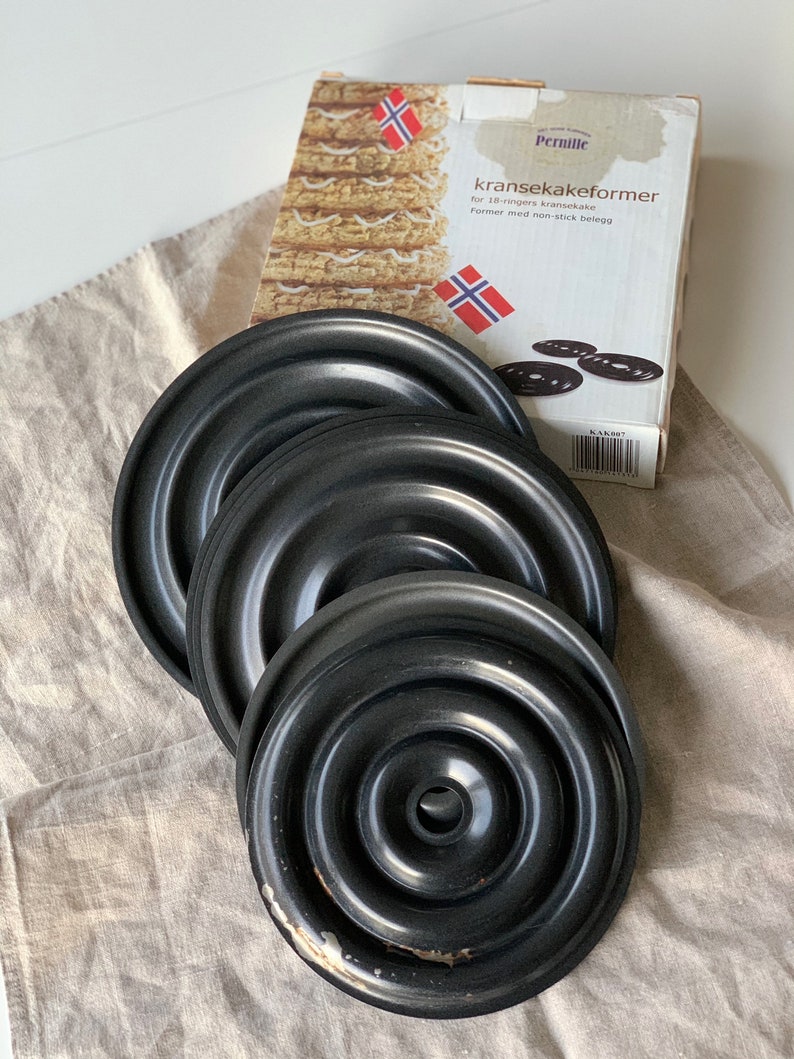 Op de afbeelding: Vier zwarte metalen taartringen voor het maken van een traditionele Noorse kransekake-taart. De ringen zijn in elkaar geschoven en hebben een antiaanbaklaag. De verpakking toont een afbeelding van een afgewerkte kransekake-taart en de tekst "kransekakeformer", wat "kransekake-vormen" betekent.