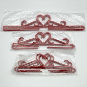 Może przedstawiać: Trzy czerwone plastikowe wieszaki z wycięciami w kształcie serca. Wieszaki są zapakowane w przezroczyste plastikowe torby.