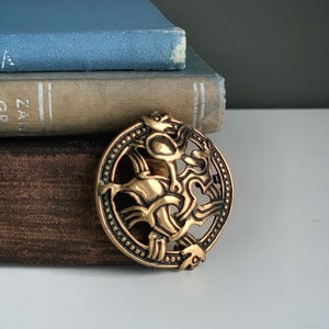 May include: A bronze-colored, circular brooch with an intricate Celtic knot design. The brooch features a detailed pattern of interwoven lines and stylized animal figures. The brooch is set against a backdrop of vintage books with blue and tan covers.