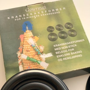 Op de afbeelding: Een set zwarte metalen ringen voor het bakken van een traditionele Noorse Kransekake-taart. De doos heeft een feestelijke illustratie van een Kransekake-taart en de tekst "Gourmet Kransekakeformer for 18-ringers Kransekake". De ringen zijn anti-aanbak voor gemakkelijk bakken en schoonmaken.