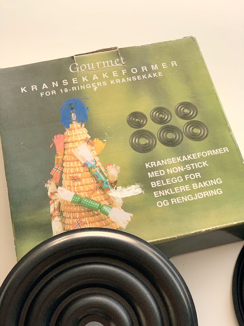 Op de afbeelding: Een set zwarte metalen ringen voor het bakken van een traditionele Noorse Kransekake-taart. De doos heeft een feestelijke illustratie van een Kransekake-taart en de tekst "Gourmet Kransekakeformer for 18-ringers Kransekake". De ringen zijn anti-aanbak voor gemakkelijk bakken en schoonmaken.