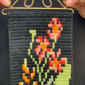 May include: A small, rectangular tapestry with a black background and a floral design. The flowers are in shades of red, orange, pink, green, and blue. The tapestry is held by a gold-colored metal hanger with decorative swirls.