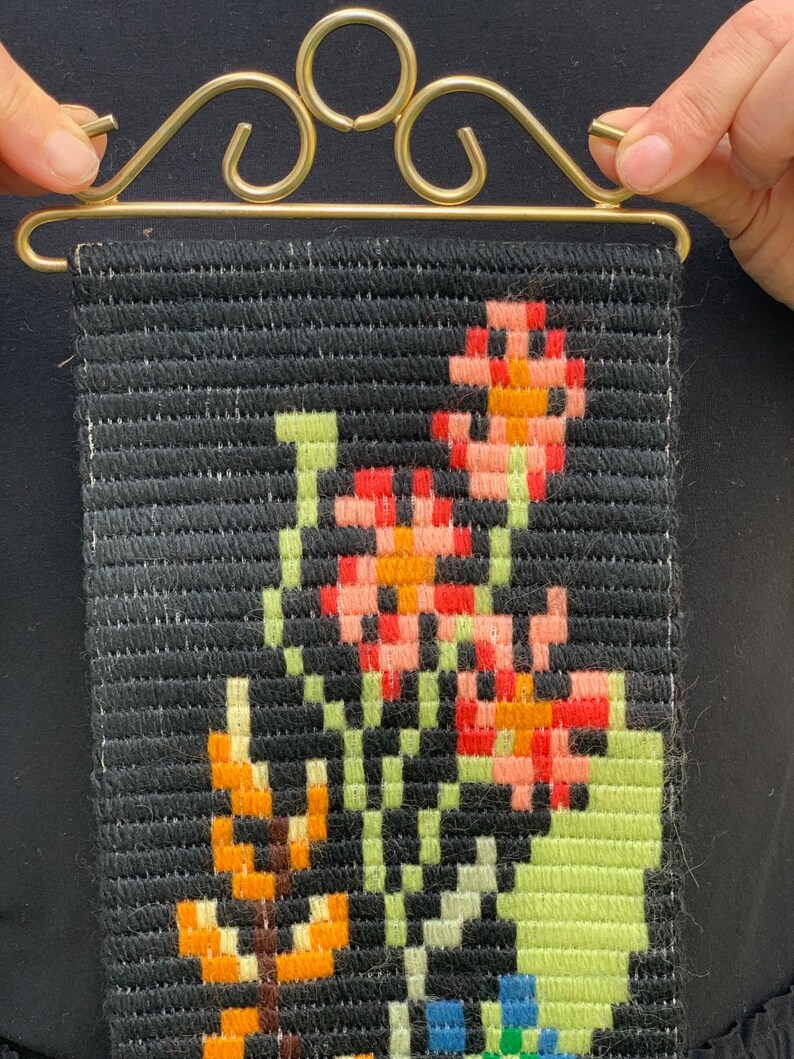 May include: A small, rectangular tapestry with a black background and a floral design. The flowers are in shades of red, orange, pink, green, and blue. The tapestry is held by a gold-colored metal hanger with decorative swirls.