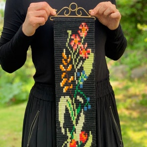 May include: A black tapestry with a floral design in orange, red, yellow, green, and blue. The tapestry is held by a gold-colored metal hanger. The floral pattern is a repeating design. The background is a black woven fabric.