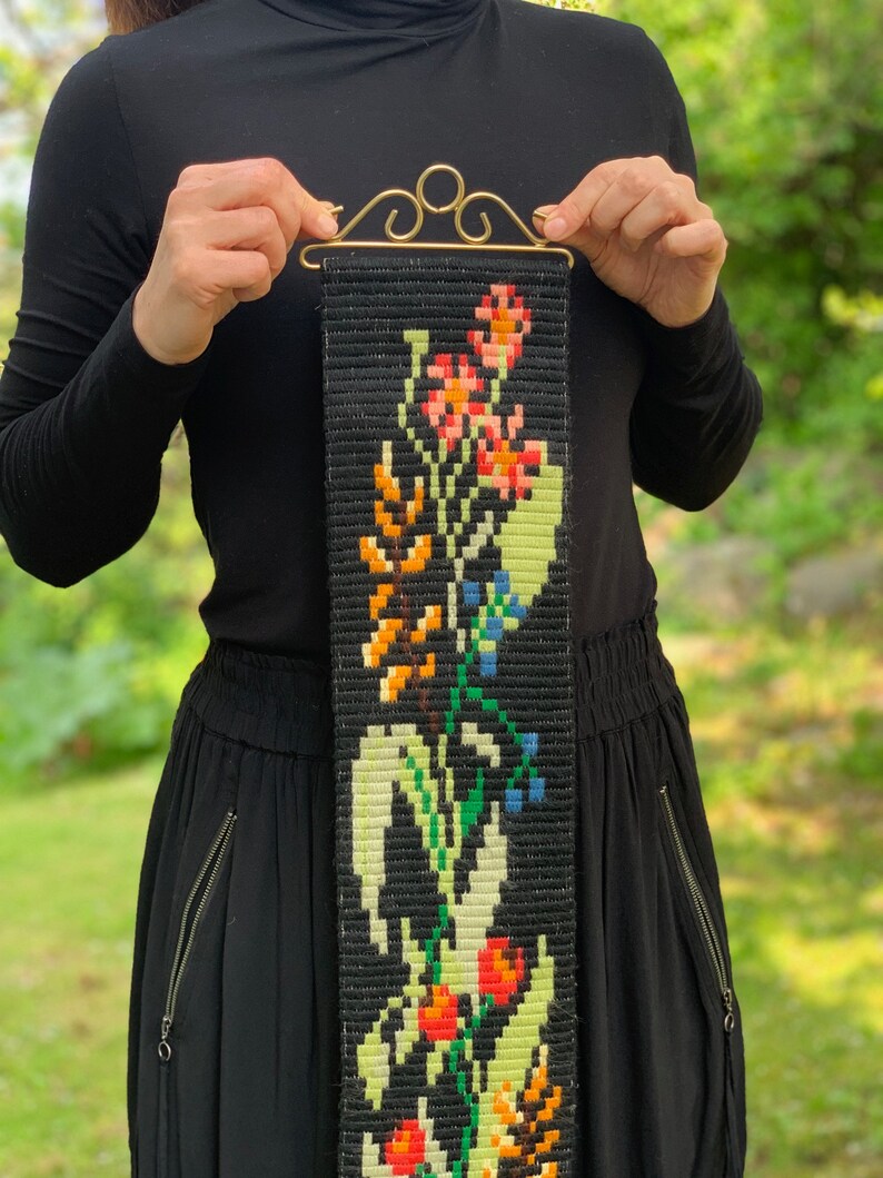 May include: A black tapestry with a floral design in orange, red, yellow, green, and blue. The tapestry is held by a gold-colored metal hanger. The floral pattern is a repeating design. The background is a black woven fabric.
