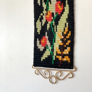 May include: A woven wall hanging with a black background and a floral design in red, green, yellow, and white. The tapestry is rectangular and has a gold-colored metal hanger at the bottom. The floral pattern includes stylized flowers and leaves.
