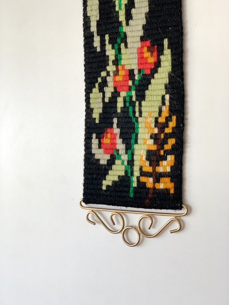 May include: A woven wall hanging with a black background and a floral design in red, green, yellow, and white. The tapestry is rectangular and has a gold-colored metal hanger at the bottom. The floral pattern includes stylized flowers and leaves.