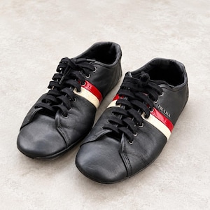 May include: A pair of black leather low-top sneakers featuring black laces and a distinctive horizontal stripe across the side in red and cream. The Prada logo is visible in silver lettering on the side of the shoe near the eyelets.