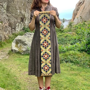 Puede incluir: Una mujer sostiene un textil rectangular con un patrón geométrico. El textil presenta un diseño repetido de formas de estrella rojas, azules y naranjas sobre un fondo amarillo. La mujer lleva un vestido marrón y zapatillas altas rojas.