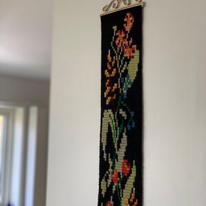 May include: A long, narrow, black tapestry with a floral design. The tapestry features colorful flowers in shades of orange, pink, yellow, and green, with blue accents. It is hung from a decorative gold-colored metal hanger.