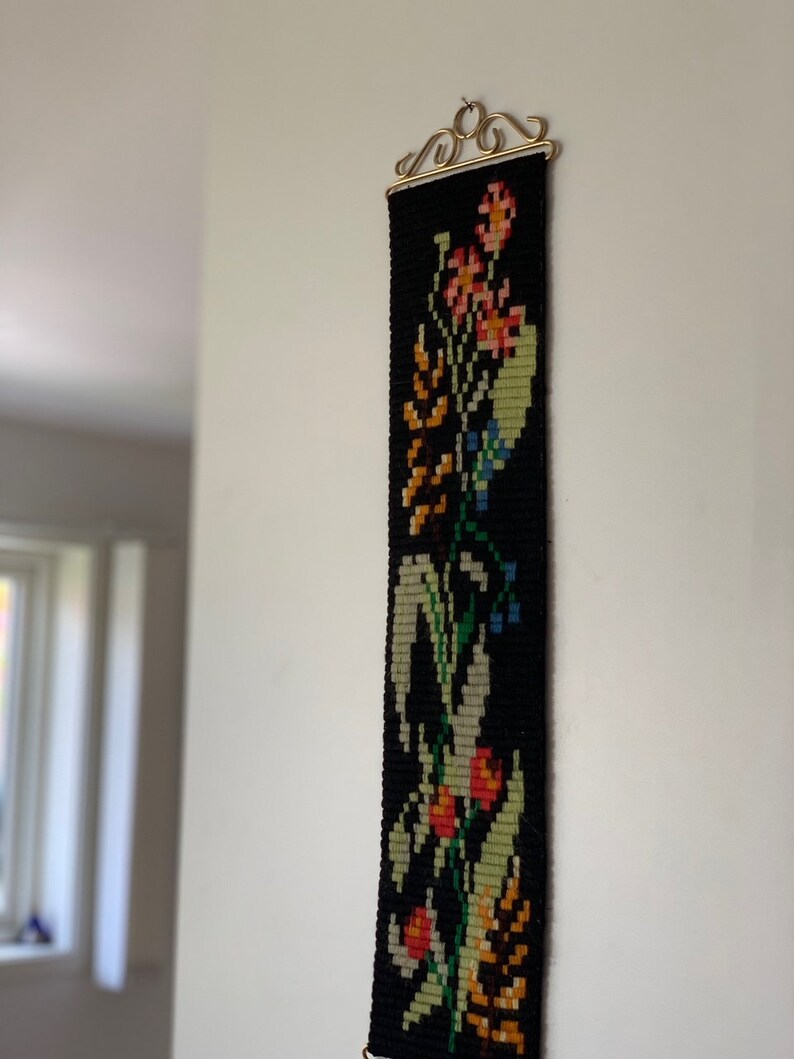 May include: A long, narrow, black tapestry with a floral design. The tapestry features colorful flowers in shades of orange, pink, yellow, and green, with blue accents. It is hung from a decorative gold-colored metal hanger.