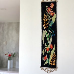 May include: A black tapestry with a floral design in shades of orange, pink, green, and yellow. The tapestry is rectangular and hangs from a decorative gold-colored metal bar. The wall hanging is mounted on a white wall.
