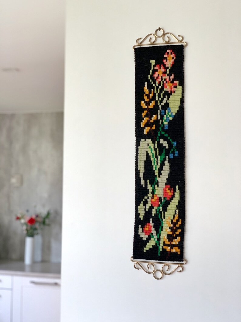 May include: A black tapestry with a floral design in shades of orange, pink, green, and yellow. The tapestry is rectangular and hangs from a decorative gold-colored metal bar. The wall hanging is mounted on a white wall.