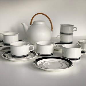 Op de afbeelding: Witte theeset met zwarte strepen. De set bevat een theepot met een geweven handvat, kopjes en schotels. De kopjes en schotels hebben een zwart streepontwerp. De theeset is op een wit oppervlak geplaatst.