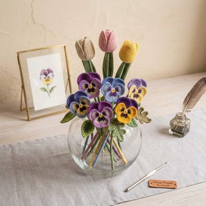 May include: A clear glass bowl filled with crocheted flowers, including three tulips in beige, pink, and yellow, and several pansies in shades of purple, blue, and yellow. The flowers have green stems and leaves. A small wooden tag reads Cottage Threads Handmade.