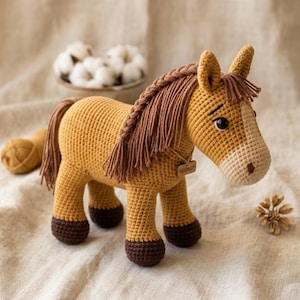 May include: A crocheted horse toy in golden brown yarn with a cream-colored muzzle, dark brown hooves, and a long, braided brown mane. The horse features black safety eyes and a small wooden tag around its neck with the word HANDMADE engraved on it.