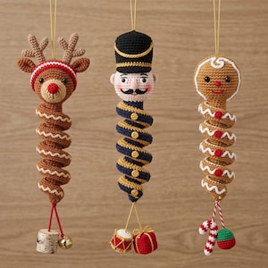 May include: Three hanging crochet ornaments featuring a reindeer with antlers, a nutcracker in a black hat, and a gingerbread person. Each figure has a long, spiralling body and is decorated with small charms like a drum, gift, and candy cane.