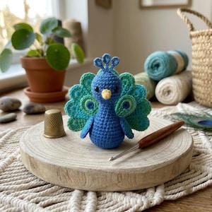 May include: A small, handmade crochet peacock figurine featuring a vibrant blue body, a yellow beak, and a fanned tail display in shades of teal and green. The peacock is positioned on a round wooden base next to a gold-toned metal thimble and a wooden crochet hook.