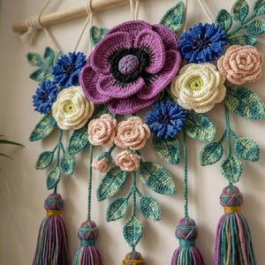 May include: A wall hanging featuring crocheted flowers in shades of purple, blue, cream, and light pink, accented with green crocheted leaves. The arrangement is suspended from a wooden dowel by white cords and includes five hanging yarn tassels.