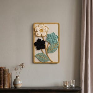 May include: A rectangular wall hanging featuring three crocheted flowers in white, black, and teal, with two teal crocheted leaves on a cream-colored fabric background, all framed by a mustard yellow crocheted border with a matching stem.