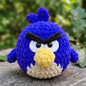 May include: A round, blue crochet plush toy featuring a textured yarn body, a yellow beak, white felt eye patches with black plastic eyes, and thick black yarn eyebrows. The character has a small tuft of blue yarn on top of its head.