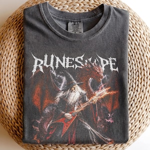 Puede incluir: Una camiseta de manga corta gris carbón con un estampado gráfico de un mago tocando una guitarra eléctrica roja con un dragón de fondo. El texto "RUNESCAPE" está impreso en una fuente blanca estilizada sobre el mago. Cuello redondo.
