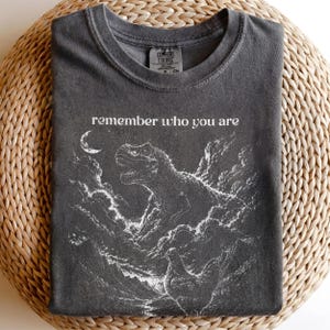 May include: A charcoal grey t-shirt featuring a white graphic print of a Tyrannosaurus Rex emerging from clouds with a crescent moon above. The text "remember who you are" is printed in white above the dinosaur. The shirt has a crew neckline.