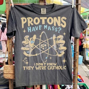 Puede incluir: Una camiseta de manga corta gris carbón con un estampado gráfico que incluye el texto "PROTONS HAVE MASS?" en beige y azul, una ilustración de un átomo sostenida por dos manos y el texto "I DIDN'T KNOW THEY WERE CATHOLIC" en letras beige.