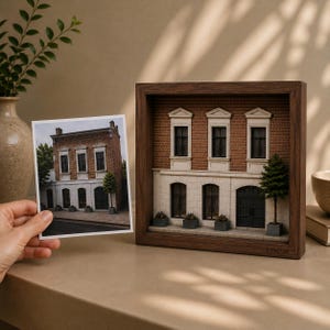 May include: A three-dimensional architectural diorama of a brick building housed in a dark wood frame, featuring white window frames, a dark door, and small potted plants. The word LOCUS is engraved on the bottom right of the wooden frame.