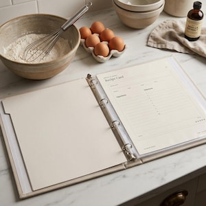 Könnte beinhalten: Ein offener Ringbuchordner mit drei Ringen, der eine cremefarbene Rezeptkartenseite mit Abschnitten für Zutaten und Anweisungen enthält, betitelt mit Write It Down Recipe Card. Der Ordner liegt auf einer weißen Oberfläche neben Mehl.