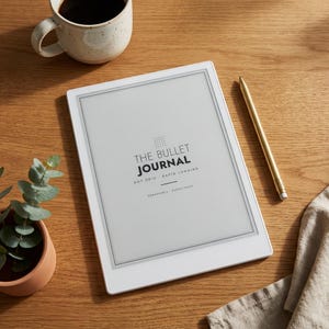 May include: A white digital tablet displaying the text THE BULLET JOURNAL DOT GRID RAPID LOGGING REMARKABLE HYPERLINKED, accompanied by a gold stylus, a speckled ceramic mug filled with dark coffee, and a small green plant in a terracotta pot.