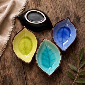 May include: A set of four leaf-shaped ceramic dishes featuring a crackle glaze finish. The dishes are coloured in lime green, turquoise, and blue, with one dish shown upside down revealing a solid black exterior and a white rimmed base.