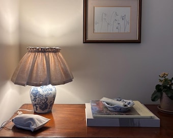 May include: A table lamp with a blue and white floral ceramic base and a pleated beige fabric shade, a framed line drawing of snowdrops, a small blue and white ceramic dish, two stacked books, and a small beige drawstring pouch.