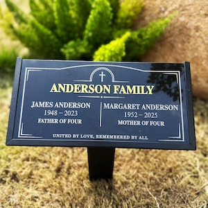May include: A rectangular black memorial plaque with a gold-coloured cross icon and the text ANDERSON FAMILY, JAMES ANDERSON 1948-2023 FATHER OF FOUR, MARGARET ANDERSON 1952-2025 MOTHER OF FOUR, and UNITED BY LOVE, REMEMBERED BY ALL, mounted on a black stake.