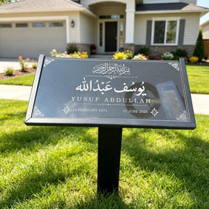 May include: A rectangular black memorial garden stake featuring white engraved text that reads Yusuf Abdullah, along with Arabic script, the dates 18 February 1971 and 17 June 2026, and decorative floral corner accents on a metal frame.