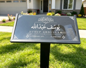 May include: A black rectangular memorial plaque on a metal stake featuring white engraved Arabic calligraphy, the name YUSUF ABDULLAH, and the dates 18 FEBRUARY 1971 and 17 JUNE 2026, accented with decorative corner floral motifs.