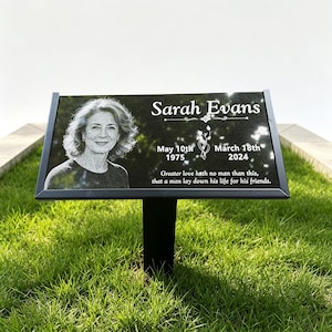 May include: A black granite memorial plaque with a metal stake, featuring an engraved portrait of Sarah Evans, her name, birth date 10th May 1975, death date 18th March 2024, and the inscription: Greater love hath no man than this, that a man lay down his life for his friends.