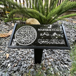 May include: A rectangular black granite memorial plaque with a black metal frame and stake. The plaque features white engraved Arabic calligraphy in a circular design, the name MARYAM YUSUF, dates 18 MARCH 1977 and 15 APRIL 2026, and Arabic text.