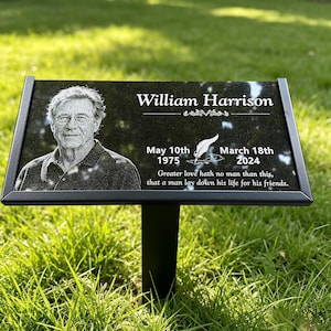Peut inclure: Une plaque commémorative rectangulaire noire sur piquet métallique, gravée du portrait de William Harrison, des dates 10 mai 1975 et 18 mars 2024, d'une plume d'écriture et du texte Greater love hath no man than this, that a man lay down.