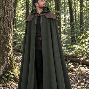 Puede incluir: Una capa con capucha de color verde oscuro que llega hasta el suelo, con detalles de cuero marrón en los hombros y un cierre de hebilla en la parte delantera. La prenda se lleva sobre una camisa negra de manga larga, pantalones negros y cinturón.