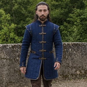 May include: A long-sleeved, dark blue quilted gambeson jacket featuring a high collar, vertical stitching, and a front closure secured by four brown leather straps with metal buckles. The garment includes shoulder pauldrons and side waist straps.