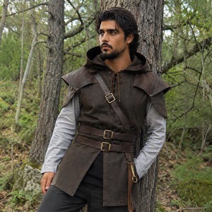 May include: A dark brown hooded tunic featuring a lace-up neckline, shoulder capes, and two adjustable leather-style belts with brass-toned buckles worn over a light gray long-sleeved shirt and dark trousers, creating a layered ensemble.