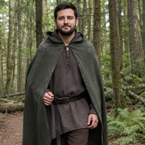 Puede incluir: Una capa con capucha de color verde oliva oscuro sobre una túnica de manga larga de color marrón oscuro con escote acordonado. Un cinturón de cuero marrón oscuro con hebilla metálica se ajusta a la cintura sobre pantalones marrones.