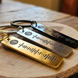 Könnte beinhalten: Drei rechteckige Metallschlüsselanhänger in Gold-, Silber- und Schwarztönen. Jeder Schlüsselanhänger besteht aus einem runden Ring und einem flachen, länglichen Metallanhänger, der mit einem Spotify-Logo und einem Schallwellenmuster graviert ist.