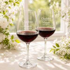 Puede incluir: Dos copas de vino de cristal transparente llenas de vino tinto. Cada copa está grabada con el texto Charlotte & Mason y la fecha April 18, 2026. Las copas presentan un tallo largo y fino y un cuenco ancho y redondeado sobre una superficie.